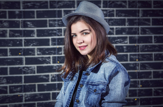 Portrait of beautiful smiling brunette teen girl, wearing jeans jacket and stylish hat, posing against dark brick wall