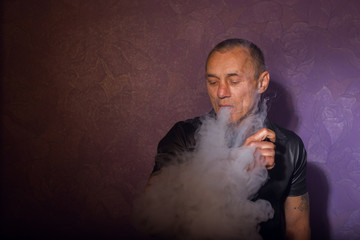 Serious tattooed man smoking vaporizer, releases clouds of vapor