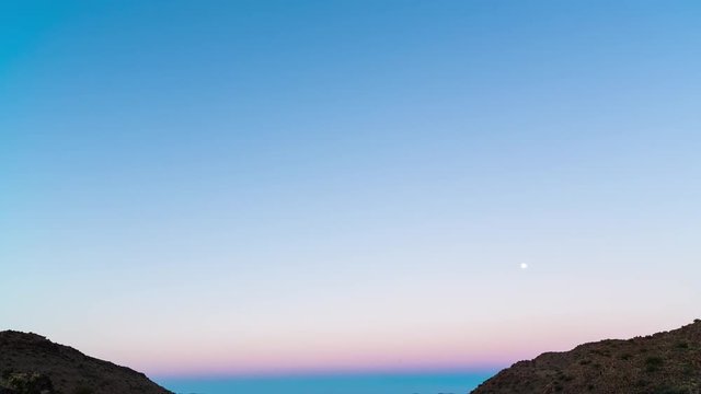 A static timelapse tilting down at dusk over vast mountain landscape with wide open plains below as the moon sets against a blue and magenta sky before sunrise