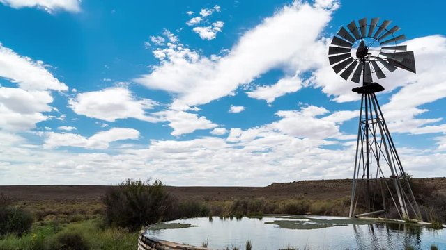 A linear midday timelapse of a windmill blowing in the wind next to and old zinc farm dam with scattered clouds against a bright blue sky in a typical Karoo landscape
