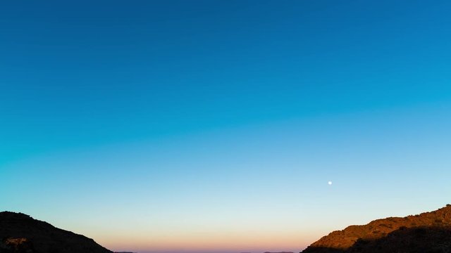 A static timelapse tilting down at sunrise over vast mountain landscape with wide open plains below as the moon sets against a blue sky and rocky hills