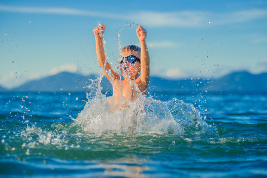 Waist portrait on a tropical beach: handsome boy in goggles jumps up and landed in the water, a lot of splashes fly apart - ready to fly like a bird, having fun