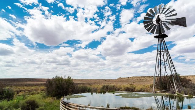 A linear midday timelapse of a windmill blowing in the wind next to and old zinc farm dam with scattered clouds against a bright blue sky in a typical Karoo landscape