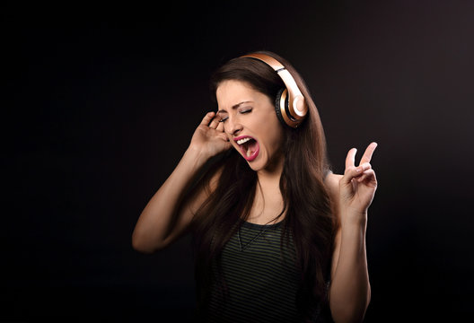 Beautiful emotional loudly singing woman listening the music in wireless headphone and hand showing v sign on dark background