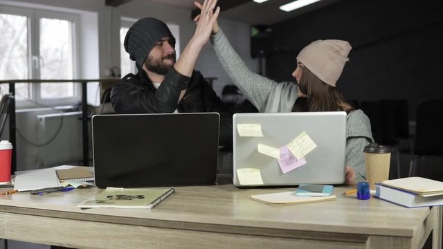 Young man and woman are working together in front of their laptops, sitting together at the table. They celebrate a successful event with a high-five. Slow motion.