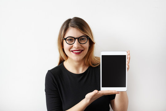 Stylish young Caucasian female in black dress and glasses looking at camera and laughing cheerfully, holding her digital tablet showing to blank tablet pc monitor. Copy space for your content