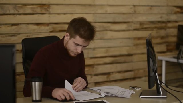 Young office worker cheking his documents in front of computer at office. Spinning a lighter. Smoker