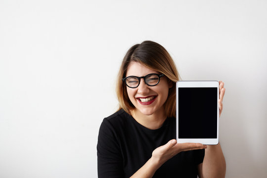 Humorous portrait of attractive young woman in glasses showing blank copy screen tablet, having funny sarcastic face expression, loud laughing meaning gloating, talking "muahaha". Copy space for text