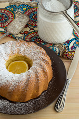 Homemade vanilla bundt cake, jar with sugar powder, sieve, towel with patterns, on a wooden background