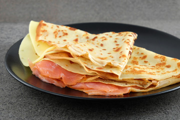 Two pancakes with salmon and cheese on black plate.