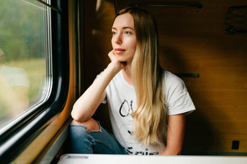 Fototapeta na wymiar Very beautiful and attractive skinny teen girl with long hair in sleeveless shirt traveling by train alone on vacation and looking at window to summer landscape. Deep mood concept.