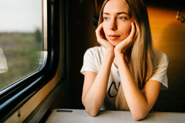 Very beautiful and attractive skinny teen girl with long hair in sleeveless shirt traveling by train alone on vacation and looking at window to summer landscape. Deep mood concept.