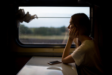 Very beautiful and attractive skinny teen girl with long hair in sleeveless shirt traveling by train alone on vacation and looking at window to summer landscape. Deep mood concept.