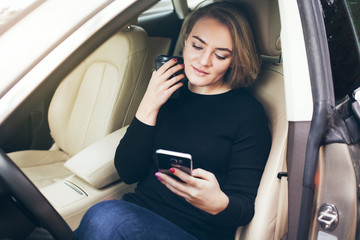Beautiful adult woman is sitting inside a car on the drivers seat, resting, drinking take away cup of coffee and looking at mobile phone using car sharing application.