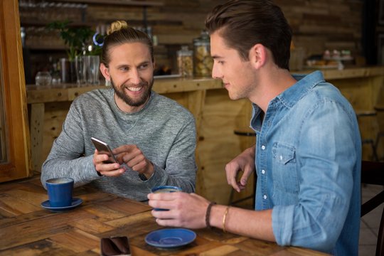 Happy Man Showing Smart Phone To Friend In Coffee Shop