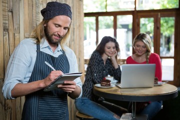 Male barista writing orders with female customers in background