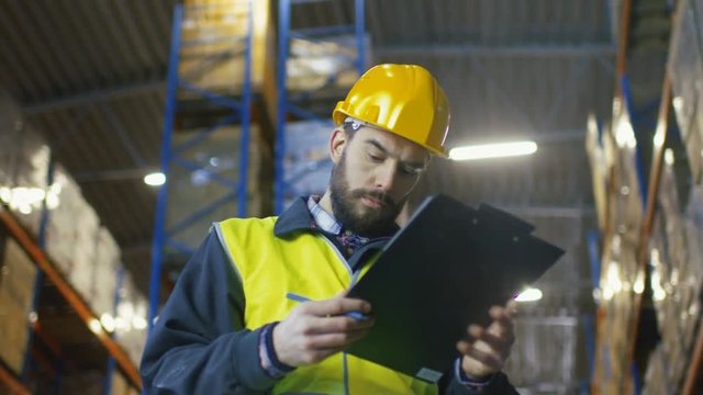  Inspector Wearing Hard Hat with Clipboard Fills in Forms in a Warehouse. He Walks Through Rows of Storage Racks with Merchandise.Shot on RED EPIC-W 8K Helium Cinema Camera.