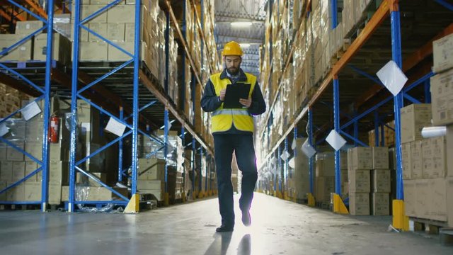 Overseer Wearing Hard Hat with Tablet Computer Counts Merchandise in Warehouse. He Walks Through Rows of Storage Racks with Merchandise. Shot on RED EPIC-W 8K Helium Cinema Camera.