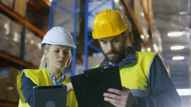  Female Supervisor with Tablet Computer and Male Surveyor with Clipboard Have Discussion in Warehouse with Big Pallet Racks in it. Shot on RED EPIC-W 8K Helium Cinema Camera.