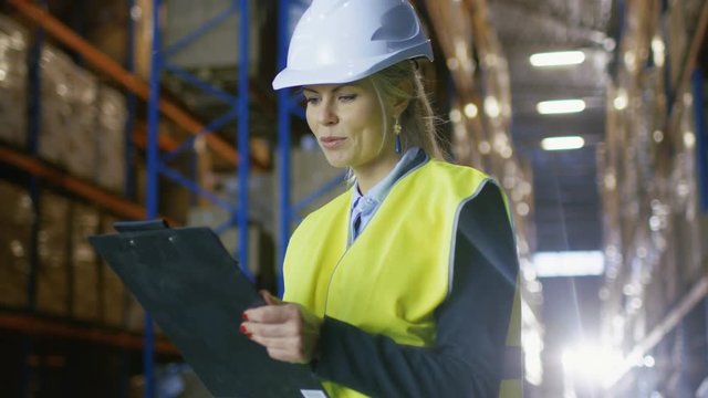 Female Controller Wearing Hard Hat and Holding Clipboard Counts Merchandise Standing in Big Warehouse with Pallet Racks in it. Shot on RED EPIC-W 8K Helium Cinema Camera.