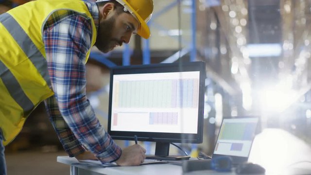 Male Inspector Fills up Spreadsheets on His Personal Computer. He's in Big Warehouse with Rows of Pallet Racks. Shot on RED EPIC-W 8K Helium Cinema Camera.