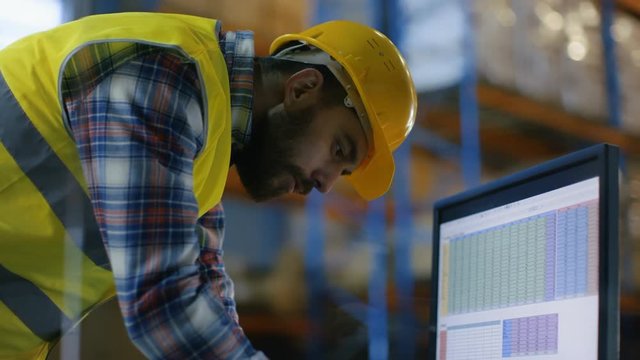Male Inspector Wearing Hard Hat Fills in Spreadsheets on His Personal Computer. He's in Big Warehouse with Rows of Pallet Racks. Shot on RED EPIC-W 8K Helium Cinema Camera.