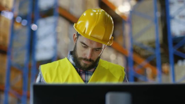 Male Inspector Wearing Hard Hat Writes in Clipboard and Consults His Personal Computer. He's in Big Warehouse with Rows of Pallet Racks. Shot on RED EPIC-W 8K Helium Cinema Camera.