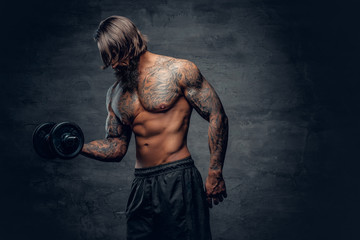 Tattooed male doing a biceps workout with dumbbell over dark grey background.
