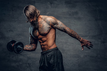 Tattooed male doing a biceps workout with dumbbell over dark grey background.