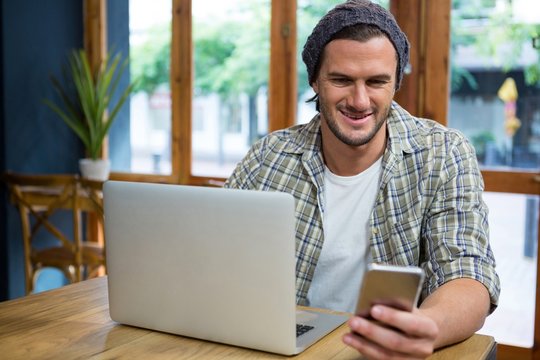 Smiling man using mobile phone and laptop in coffee shop