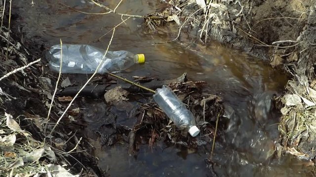 Empty plastic bottles in spring creek. Environmental pollution concept. Water flow among dirt, old papers and froth moving in stream.