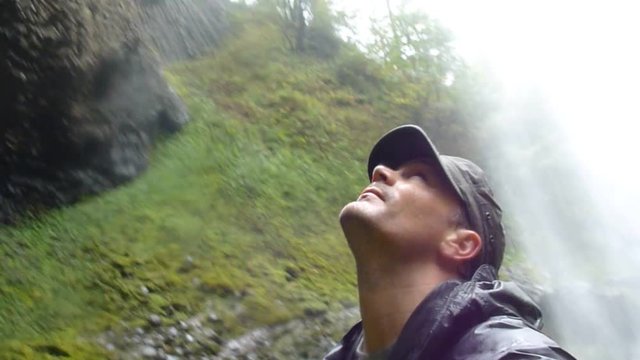 Man under large waterfall in Oregon on rainy day waives hand to camera and looks up.
