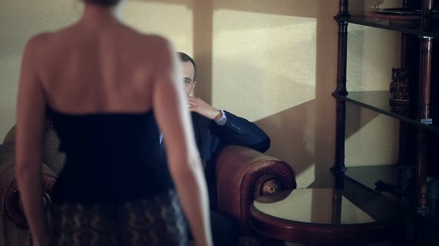 A girl in lacy underwear comes to a man in a suit who expects her sitting in a brutal chair with a glass of cognac