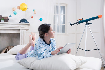 Cute girl in pajamas lying on bed with digital tablet and looking at telescope - Powered by Adobe