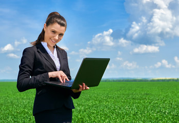 Business woman in green grass field outdoor work on laptop. Young girl dressed in black suit. Beautiful spring landscape with cloudy sky, bright sunny day