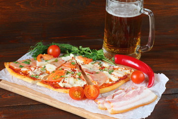 Pizza with smoked meat and a mug of beer are on a dark wooden background.