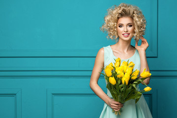 Beautiful blonde girl in the blue dress with flowers tulips in hands on a turquoise background