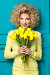 Beautiful blonde girl in yellow shirt with flowers tulips in hands on a turquoise background