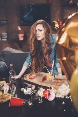 Depressed young woman with hangover leaning at messy table after party