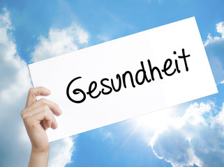 Gesundheit (Health in German)  Sign on white paper. Man Hand Holding Paper with text. Isolated on sky background.