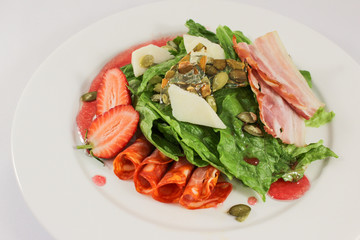 Mix of hot sausages with bacon on a cushion of lettuce leaves, in a duet with parmesan and strawberries, garnished with pumpkin seeds, dressed with raspberry sauce