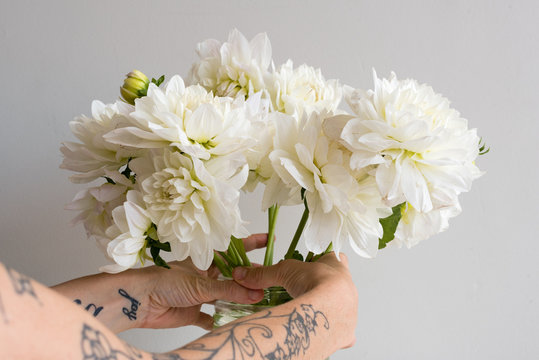 Woman's arms with partly visible tattoos arranging white dahlias in glass vase against neutral background (selective focus)