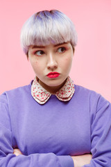 Fashion close-up portrait of angry beautiful dollish girl with short light violet hair wearing lilac sweater over pink background.