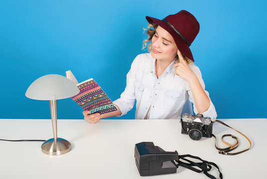 Happy young hipster girl photographer sitting at the table,looking to notebook,equipment.woman in red hat,blue background,happy face,girl with camera,curly hair,attractive woman,traveler girl 