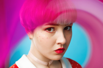 Close up artistic conceptual portrait of beautiful dollish girl with short light violet hair wearing red dress over blue background. Blurry forefront.