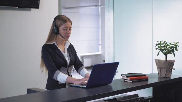 Young professional woman receptionist standing at reception desk talking with client check information in computer. Smiling female with long blond hair using hands free in office. Friendly worker in