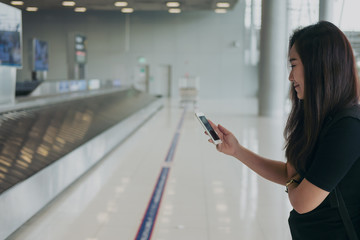 A beautiful Asian woman using mobile phone with feeling happy and smiley face , standing and waiting for baggage claim in the airport