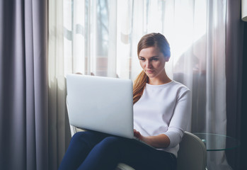 Pretty woman in modern hotel room sitting on armchair and using laptop