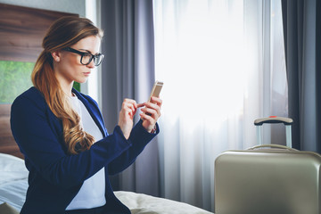 Business woman with suitcase in modern hotel room using laptop and smartphone