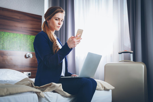 Business woman with suitcase in modern hotel room using laptop and smartphone